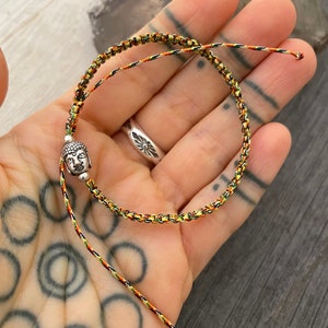 May include: A braided cord bracelet featuring a small silver-toned Buddha head charm. The cord is woven with a multi-colored pattern of green, yellow, and red threads. The bracelet has two loose ends and a sliding knot mechanism for adjustment.