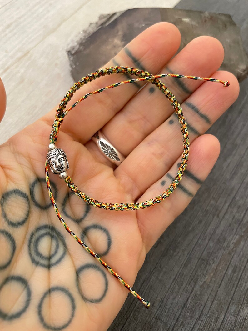 May include: A braided cord bracelet featuring a small silver-toned Buddha head charm. The cord is woven with a multi-colored pattern of green, yellow, and red threads. The bracelet has two loose ends and a sliding knot mechanism for adjustment.