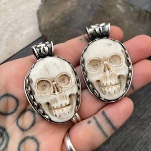 May include: Two white bone skull pendants with silver accents. The skulls are carved in detail and have a realistic look. The pendants are oval shaped and have a silver bail at the top.