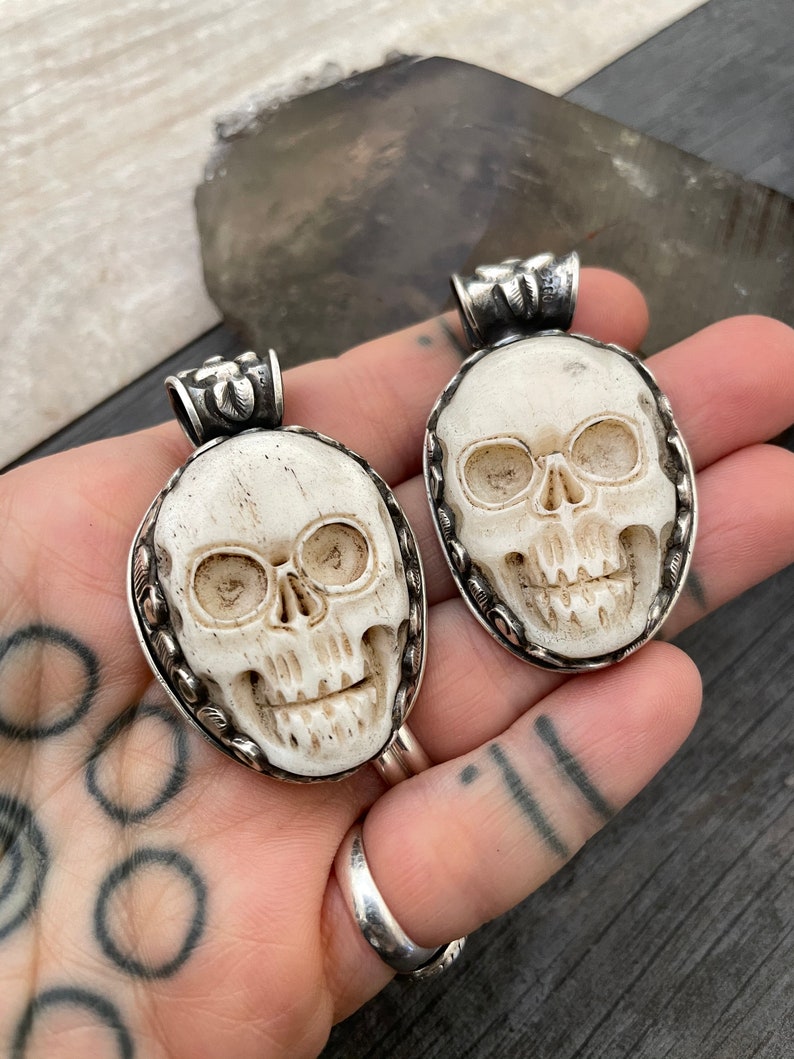 May include: Two white bone skull pendants with silver accents. The skulls are carved in detail and have a realistic look. The pendants are oval shaped and have a silver bail at the top.