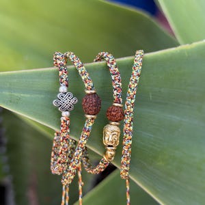 May include: A set of three braided bracelets made from multicolored thread in shades of yellow, red, green, and blue. One bracelet features a silver-toned eternal knot charm, while the others include brown textured beads and a gold-toned Buddha head.