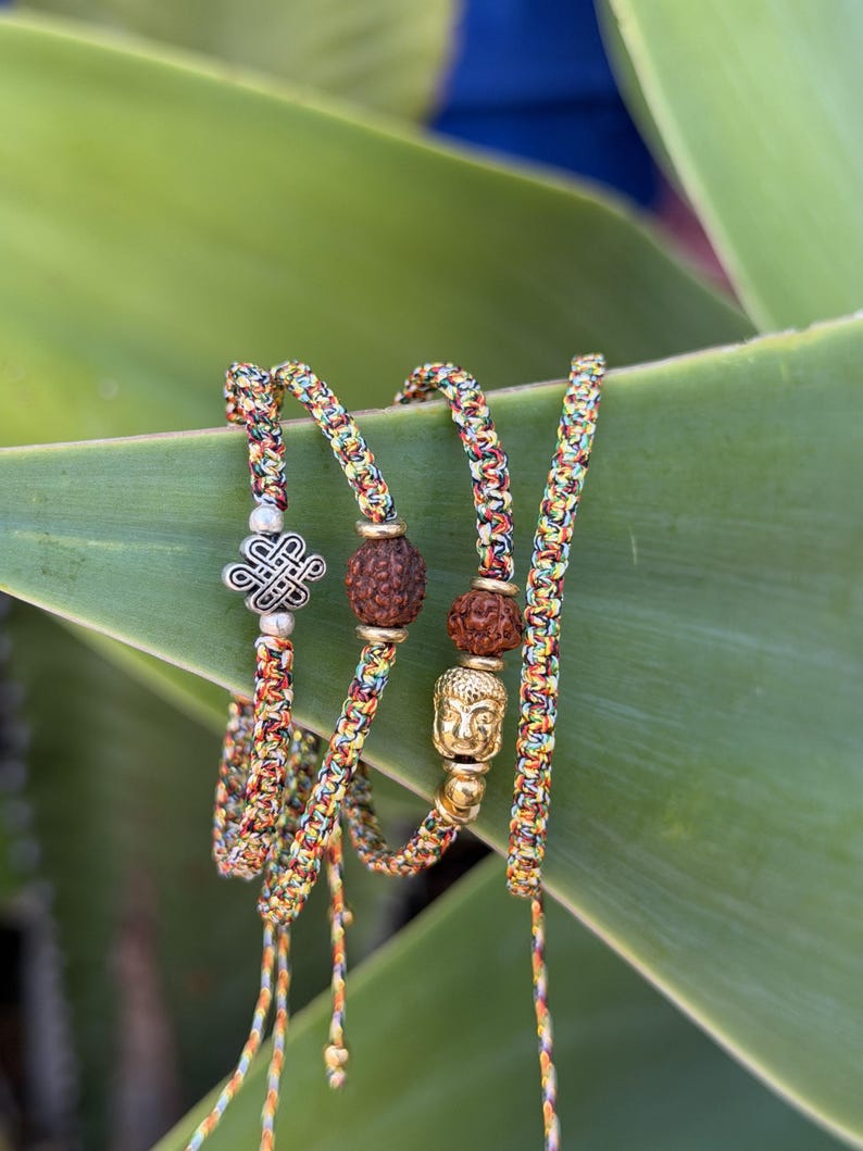 May include: A set of three braided bracelets made from multicolored thread in shades of yellow, red, green, and blue. One bracelet features a silver-toned eternal knot charm, while the others include brown textured beads and a gold-toned Buddha head.