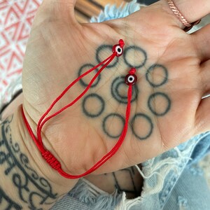 K&ouml;nnte beinhalten: Rotes Armband mit zwei b&ouml;sen Augen-Anh&auml;ngern. Die Anh&auml;nger sind blau und wei&szlig; mit einem roten Zentrum.