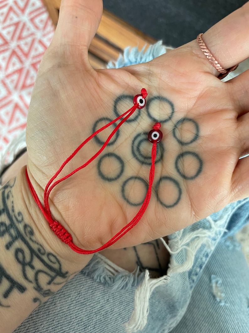 K&ouml;nnte beinhalten: Rotes Armband mit zwei b&ouml;sen Augen-Anh&auml;ngern. Die Anh&auml;nger sind blau und wei&szlig; mit einem roten Zentrum.