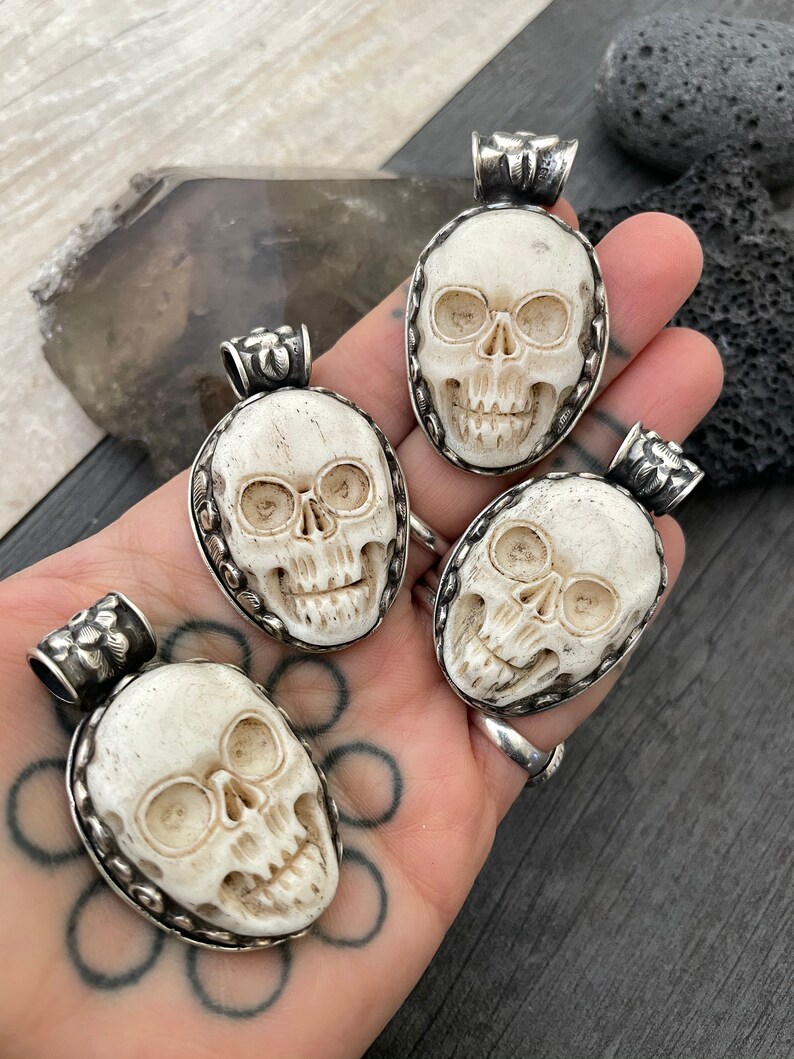 May include: Four white bone skull pendants with silver accents. Each pendant is carved with a detailed skull design and has a silver bail for hanging.