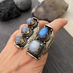 May include: A silver ring with three moonstone gems set in a curved band. The ring is decorated with intricate floral designs and has a textured finish.
