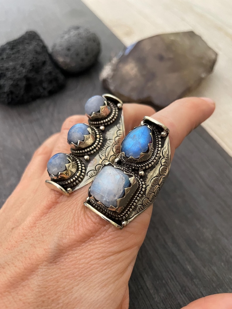 May include: A silver ring with three moonstone gems set in a curved band. The ring is decorated with intricate floral designs and has a textured finish.
