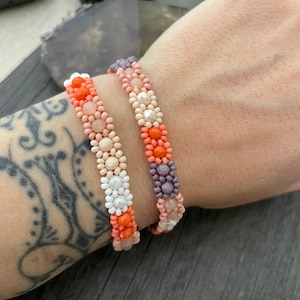 Könnte beinhalten: Zwei Perlenarmbänder mit einem mehrfarbigen Muster aus rosa, orange, weißen und violetten Perlen. Die Armbänder sind auf dem Handgelenk einer Person übereinander gelegt.