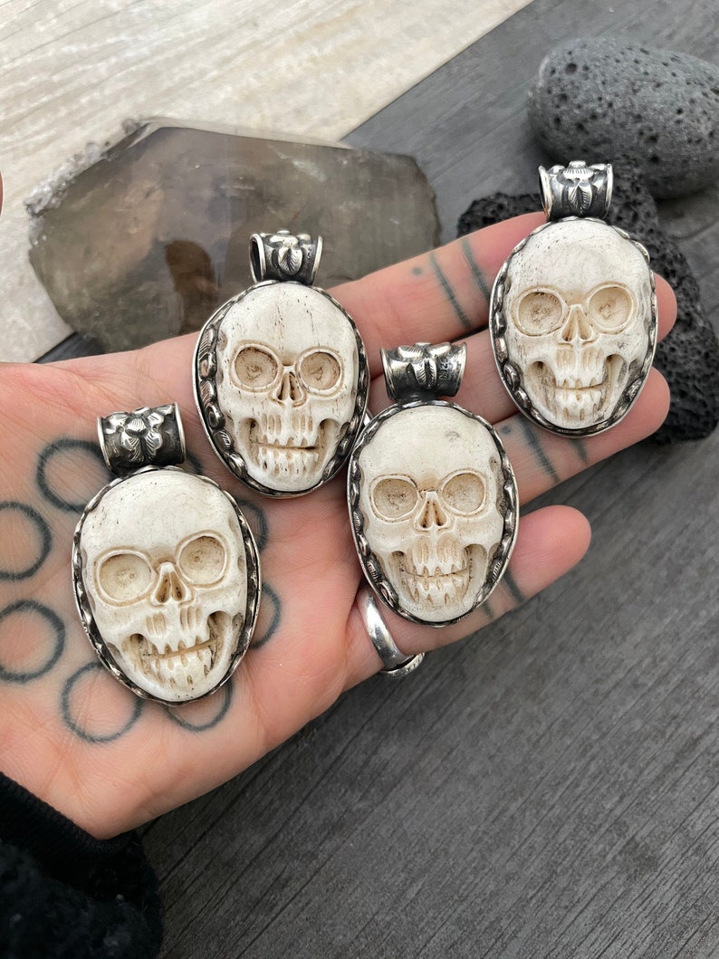 May include: Four white bone skull pendants with silver accents. Each pendant features a detailed carving of a skull with eye sockets, a nose, and a mouth.