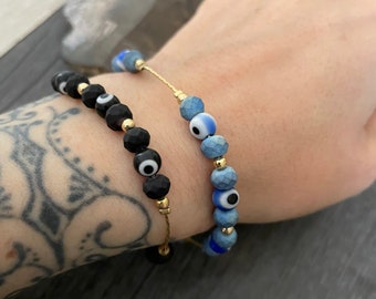 May include: Two bracelets with evil eye beads. One bracelet has black beads with gold accents and the other has blue beads with gold accents.