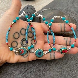 Puede incluir: Tres pulseras de cuentas turquesa con cierres plateados y dijes redondos decorativos. Los dijes son de metal y presentan piedras turquesa, rojas y azules.