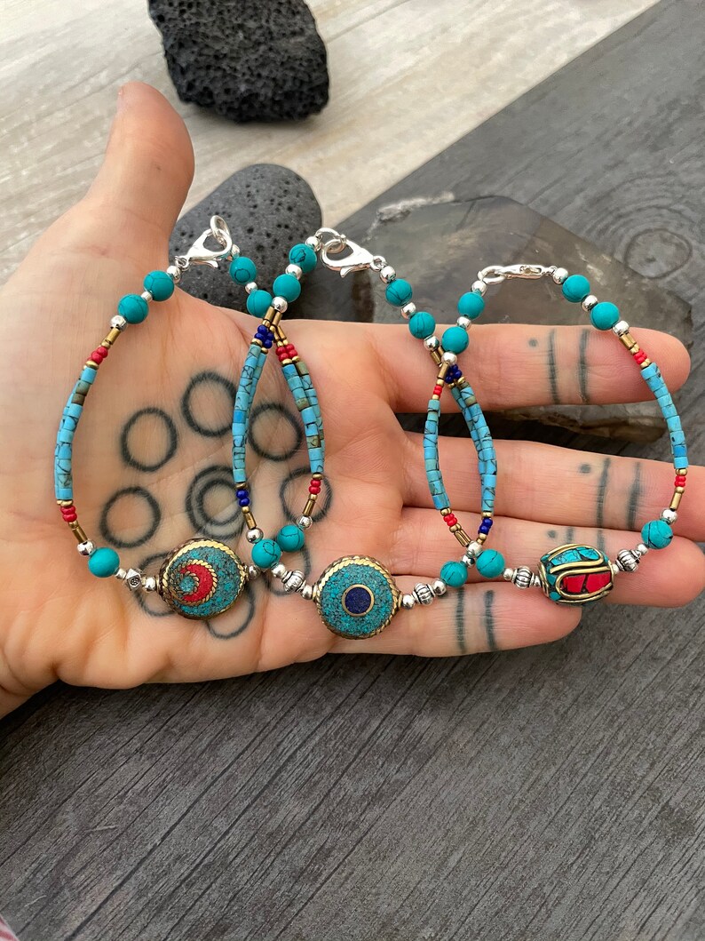 Puede incluir: Tres pulseras de cuentas turquesa con cierres plateados y dijes redondos decorativos. Los dijes son de metal y presentan piedras turquesa, rojas y azules.