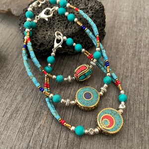 Puede incluir: Un collar de cuentas de turquesa y plata en capas con tres colgantes decorativos. Los colgantes est&aacute;n hechos de lat&oacute;n y presentan piedras de turquesa, coral rojo y lapisl&aacute;zuli.
