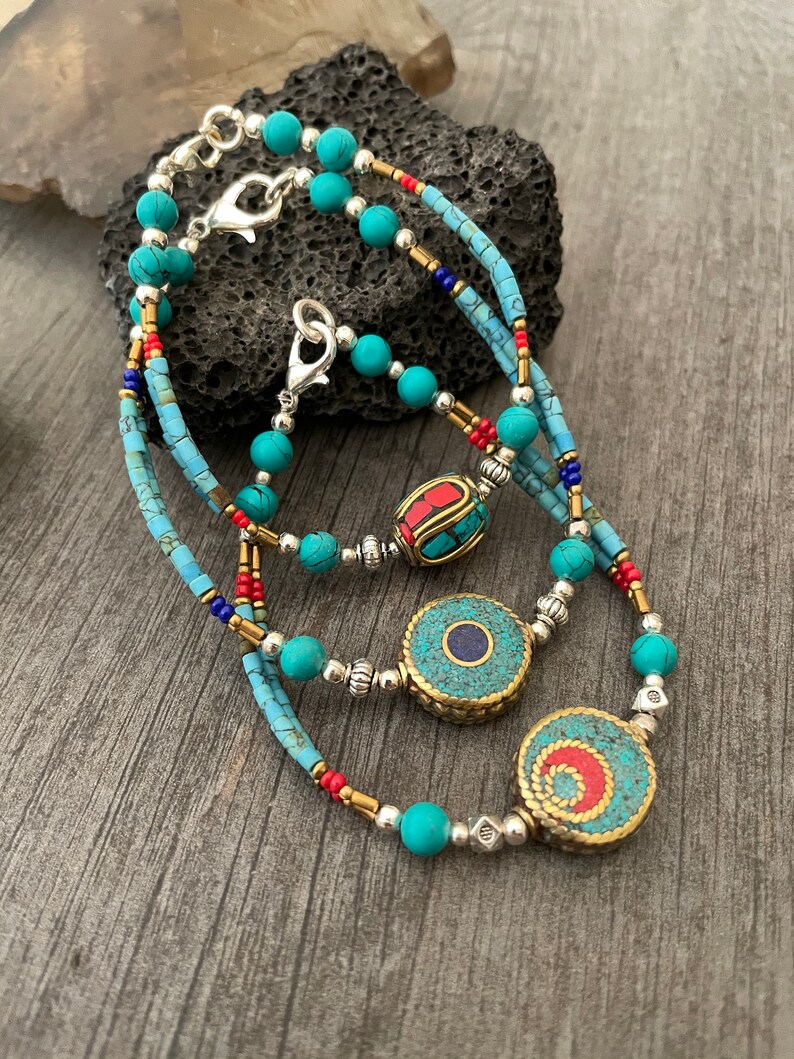 Puede incluir: Un collar de cuentas de turquesa y plata en capas con tres colgantes decorativos. Los colgantes est&aacute;n hechos de lat&oacute;n y presentan piedras de turquesa, coral rojo y lapisl&aacute;zuli.
