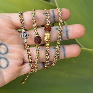 May include: Four braided bracelets made of multicolored thread in shades of yellow, red, and green. The bracelets feature metallic charms including a silver eternal knot, a gold-toned Buddha head, and two brown textured rudraksha beads.