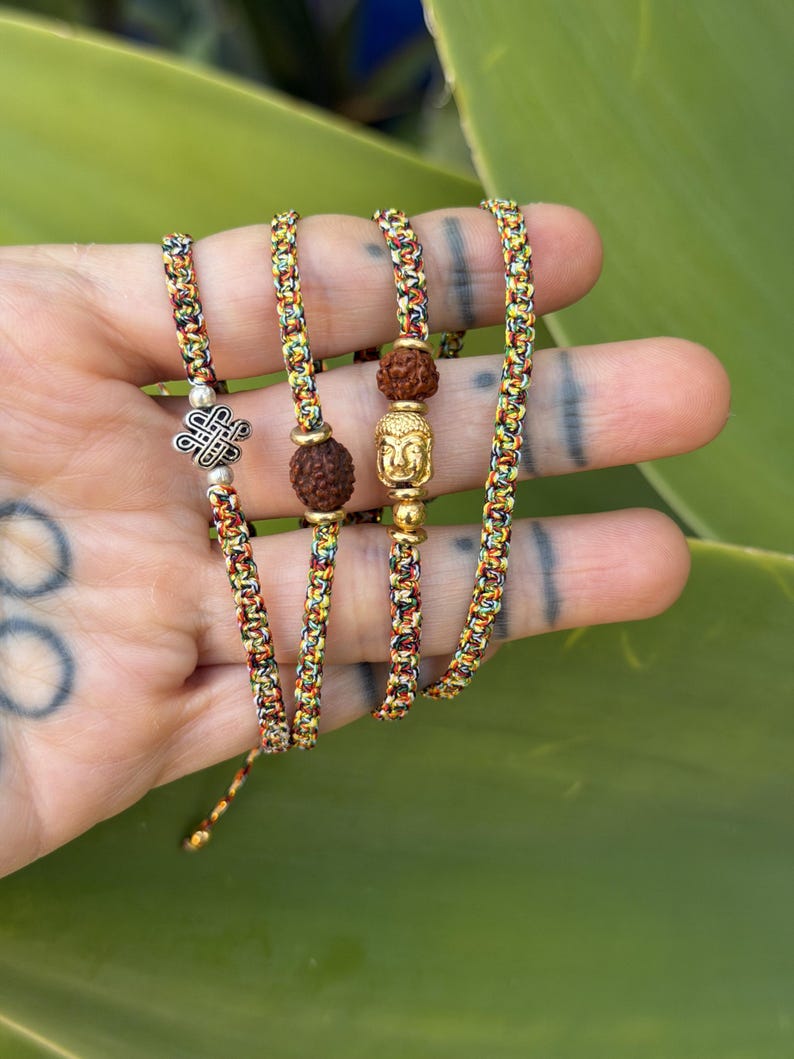 May include: Four braided bracelets made of multicolored thread in shades of yellow, red, and green. The bracelets feature metallic charms including a silver eternal knot, a gold-toned Buddha head, and two brown textured rudraksha beads.