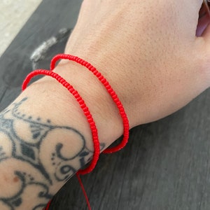 May include: Two red beaded bracelets on a person's wrist. The bracelets are made of small, round beads and are tied together with a red string.