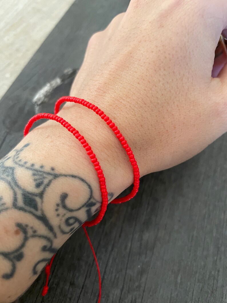 May include: Two red beaded bracelets on a person's wrist. The bracelets are made of small, round beads and are tied together with a red string.