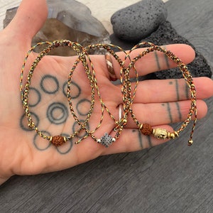 May include: Three adjustable bracelets made of braided cord in black, red, and yellow. The first features a brown rudraksha bead, the second a silver-toned mystic knot charm, and the third a gold-toned Buddha head bead with small metallic accents.