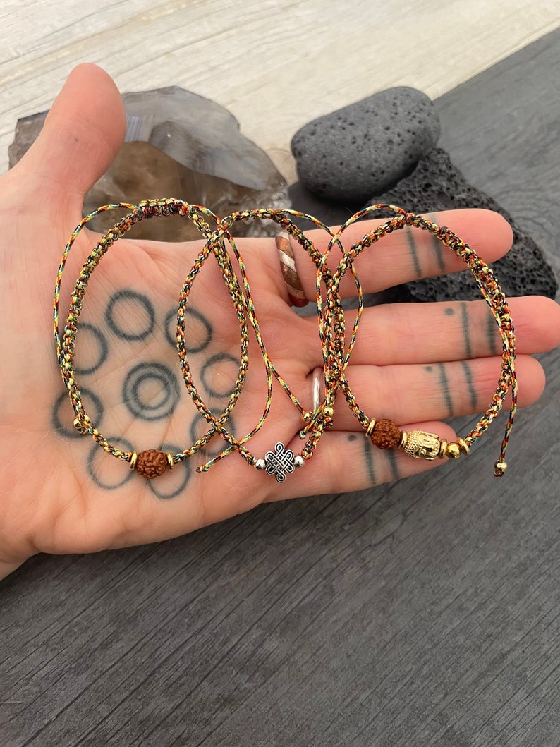 May include: Three adjustable bracelets made of braided cord in black, red, and yellow. The first features a brown rudraksha bead, the second a silver-toned mystic knot charm, and the third a gold-toned Buddha head bead with small metallic accents.