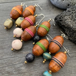 May include: A collection of handmade earrings featuring wooden acorn-shaped beads wrapped with colorful thread. The earrings have various accent beads in shades of blue, pink, and green. The earrings are displayed on a wooden surface.