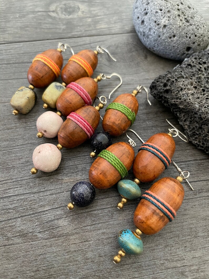 May include: A collection of handmade earrings featuring wooden acorn-shaped beads wrapped with colorful thread. The earrings have various accent beads in shades of blue, pink, and green. The earrings are displayed on a wooden surface.