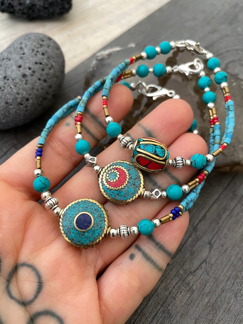 Puede incluir: Tres pulseras de cuentas turquesa y plata con diferentes piedras centrales de colores. Las piedras centrales son redondas y presentan dise&ntilde;os intrincados con piedras turquesa, rojas y azules.