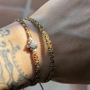 May include: Two braided bracelets made of multicolored thread in shades of yellow, green, red, and black. One bracelet features a central silver-toned metal charm shaped like an intricate knot, flanked by two small silver-toned spherical beads.