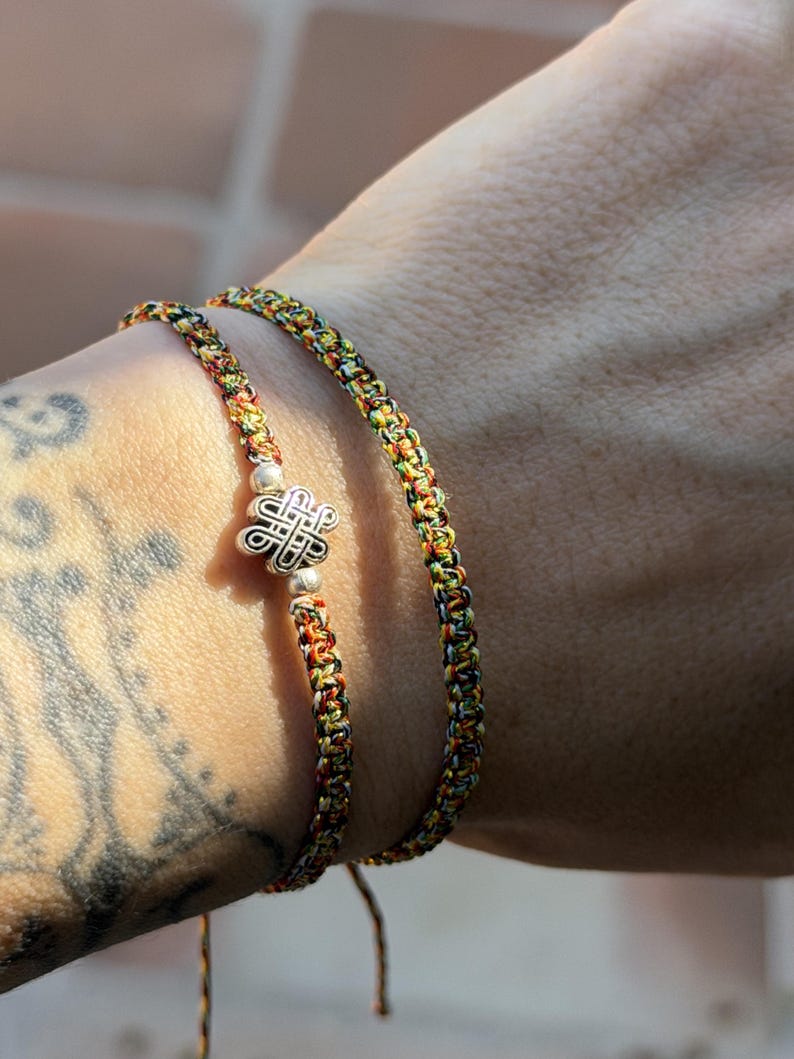 May include: Two braided bracelets made of multicolored thread in shades of yellow, green, red, and black. One bracelet features a central silver-toned metal charm shaped like an intricate knot, flanked by two small silver-toned spherical beads.