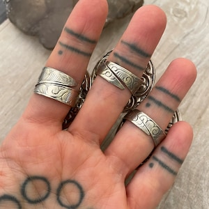 May include: Three silver rings with intricate designs on a hand with a tattoo. The rings are worn on the index, middle, and ring fingers.