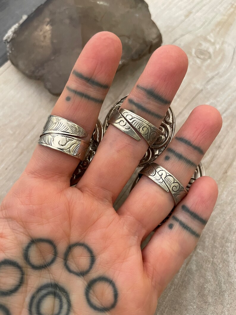 May include: Three silver rings with intricate designs on a hand with a tattoo. The rings are worn on the index, middle, and ring fingers.