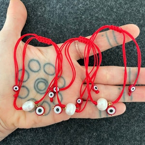 K&ouml;nnte beinhalten: Sechs rote Armb&auml;nder mit einer wei&szlig;en Perle und einem blauen Gl&uuml;cksbringer-Auge. Die Armb&auml;nder sind verknotet und liegen auf einer Hand.