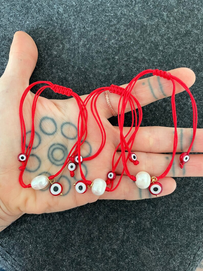 K&ouml;nnte beinhalten: Sechs rote Armb&auml;nder mit einer wei&szlig;en Perle und einem blauen Gl&uuml;cksbringer-Auge. Die Armb&auml;nder sind verknotet und liegen auf einer Hand.