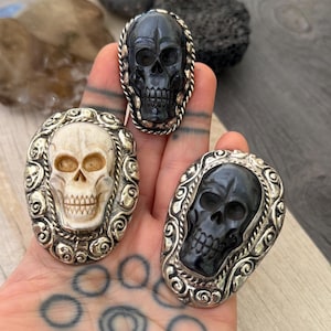 Puede incluir: Tres anillos de calavera. Los anillos presentan tallas detalladas de calaveras en negro y blanquecino, engastadas en marcos plateados ornamentados. Los anillos son de forma ovalada, con intrincados adornos alrededor de las calaveras.