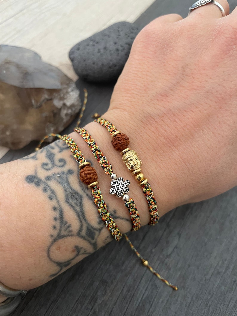 May include: Three braided bracelets featuring multicolored threads in yellow, green, and red. One bracelet includes a brown textured bead, another a silver-toned eternal knot charm, and the third a gold-toned Buddha head bead with a brown bead.