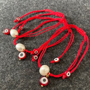K&ouml;nnte beinhalten: Set aus roten Schnurarmb&auml;ndern mit wei&szlig;en Perlen und b&ouml;sen Augen-Anh&auml;ngern. Die Anh&auml;nger sind rot mit einem wei&szlig;en Auge und goldenen Akzenten.