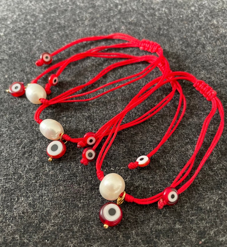 K&ouml;nnte beinhalten: Set aus roten Schnurarmb&auml;ndern mit wei&szlig;en Perlen und b&ouml;sen Augen-Anh&auml;ngern. Die Anh&auml;nger sind rot mit einem wei&szlig;en Auge und goldenen Akzenten.