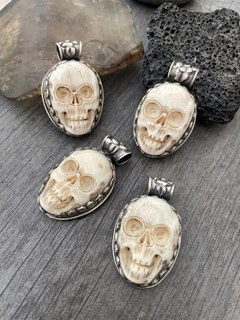May include: Four white bone skull pendants with silver accents. Each pendant is carved with a detailed skull design and has a silver bail for hanging.
