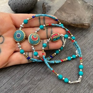 Puede incluir: Un collar de cuentas turquesa y plata con tres dijes decorativos. Los dijes est&aacute;n hechos de lat&oacute;n y presentan dise&ntilde;os intrincados con acentos turquesa y rojo.