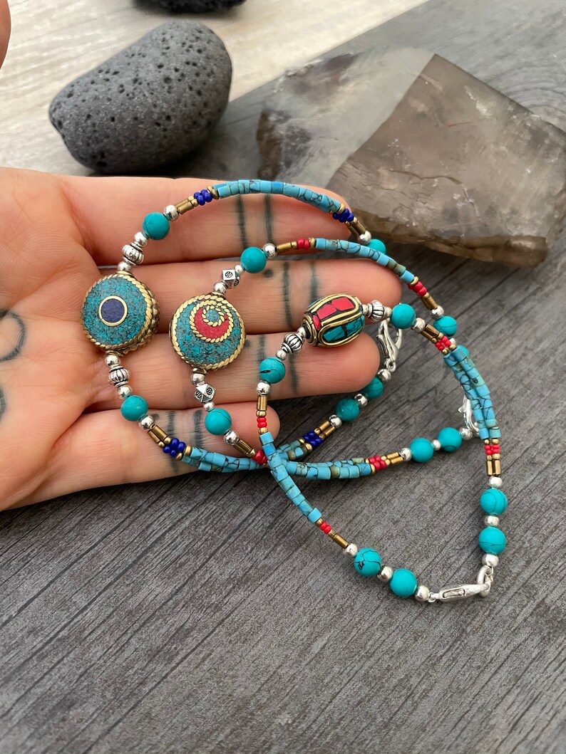 Puede incluir: Un collar de cuentas turquesa y plata con tres dijes decorativos. Los dijes est&aacute;n hechos de lat&oacute;n y presentan dise&ntilde;os intrincados con acentos turquesa y rojo.