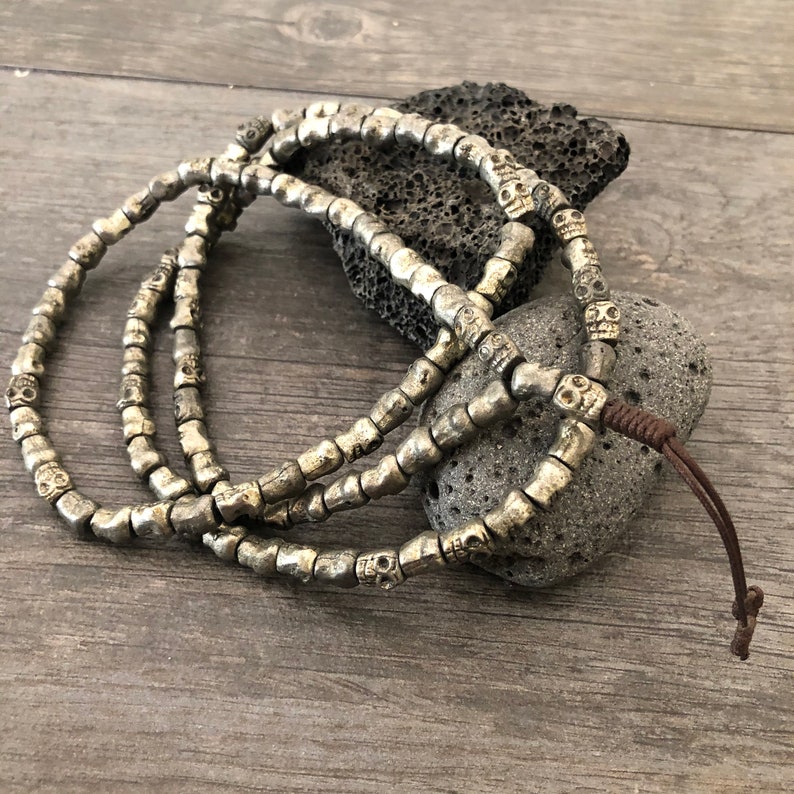 May include: A silver skull bead necklace with a brown leather cord. The necklace is made of small, round beads with a skull design.