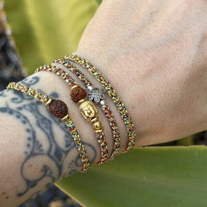 May include: Three braided bracelets made of multicolored cord in shades of yellow, orange, and green. One bracelet features a brown rudraksha bead, another includes a gold-toned Buddha charm, and the third displays a silver-toned knot charm.