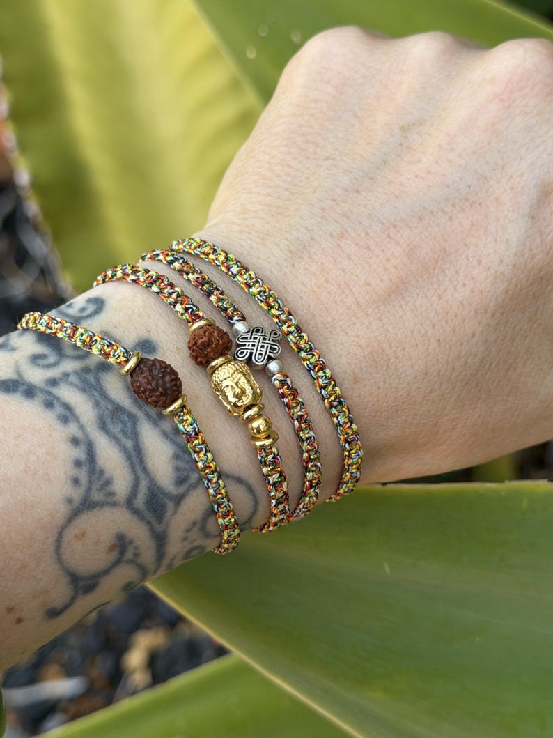 May include: Three braided bracelets made of multicolored cord in shades of yellow, orange, and green. One bracelet features a brown rudraksha bead, another includes a gold-toned Buddha charm, and the third displays a silver-toned knot charm.