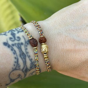 May include: Two braided bracelets made of multicolored thread featuring brown textured beads and gold-toned accents. One bracelet includes a small gold-toned Buddha head charm, while the other features a single brown textured bead.