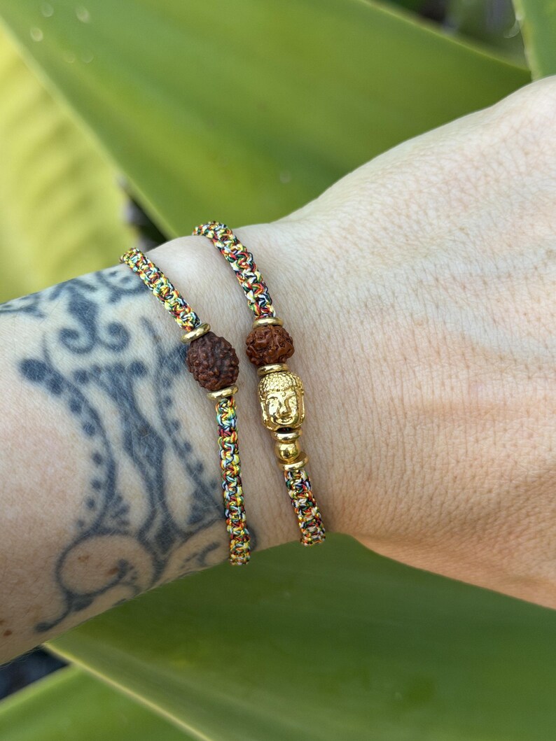 May include: Two braided bracelets made of multicolored thread featuring brown textured beads and gold-toned accents. One bracelet includes a small gold-toned Buddha head charm, while the other features a single brown textured bead.