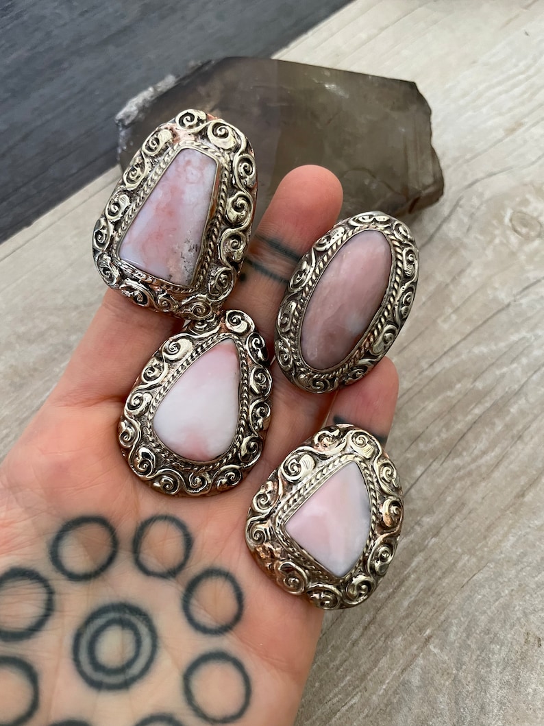 May include: Four silver rings with pink opal stones set in ornate silver settings. The rings are on a hand with a tattoo.