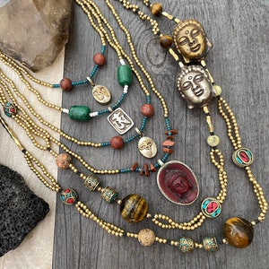 May include: A collection of layered necklaces with various beads and pendants. The necklaces feature gold-colored beads, tiger's eye beads, and pendants with Buddha faces and religious symbols. The color palette includes gold, brown, red, green, and turquoise.