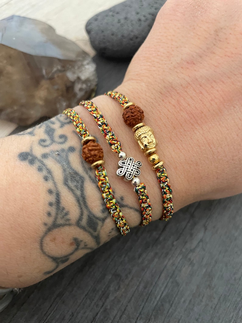 May include: Three braided bracelets featuring multicolored cord in yellow, red, green, and black. The bracelets include a brown textured bead, a gold-toned Buddha head charm, and a silver-toned eternal knot charm with small metallic spacer beads.