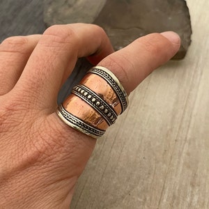 May include: A wide, copper-colored ring with silver-colored braided and beaded details. The ring is worn on a finger, showcasing its intricate design and contrasting metal tones. The ring's design is bold and eye-catching.