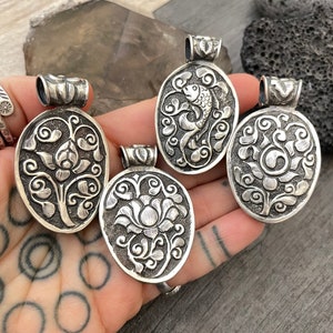 May include: Four silver pendants featuring intricate floral and fish designs. The pendants are oval shaped and have a detailed, textured surface.